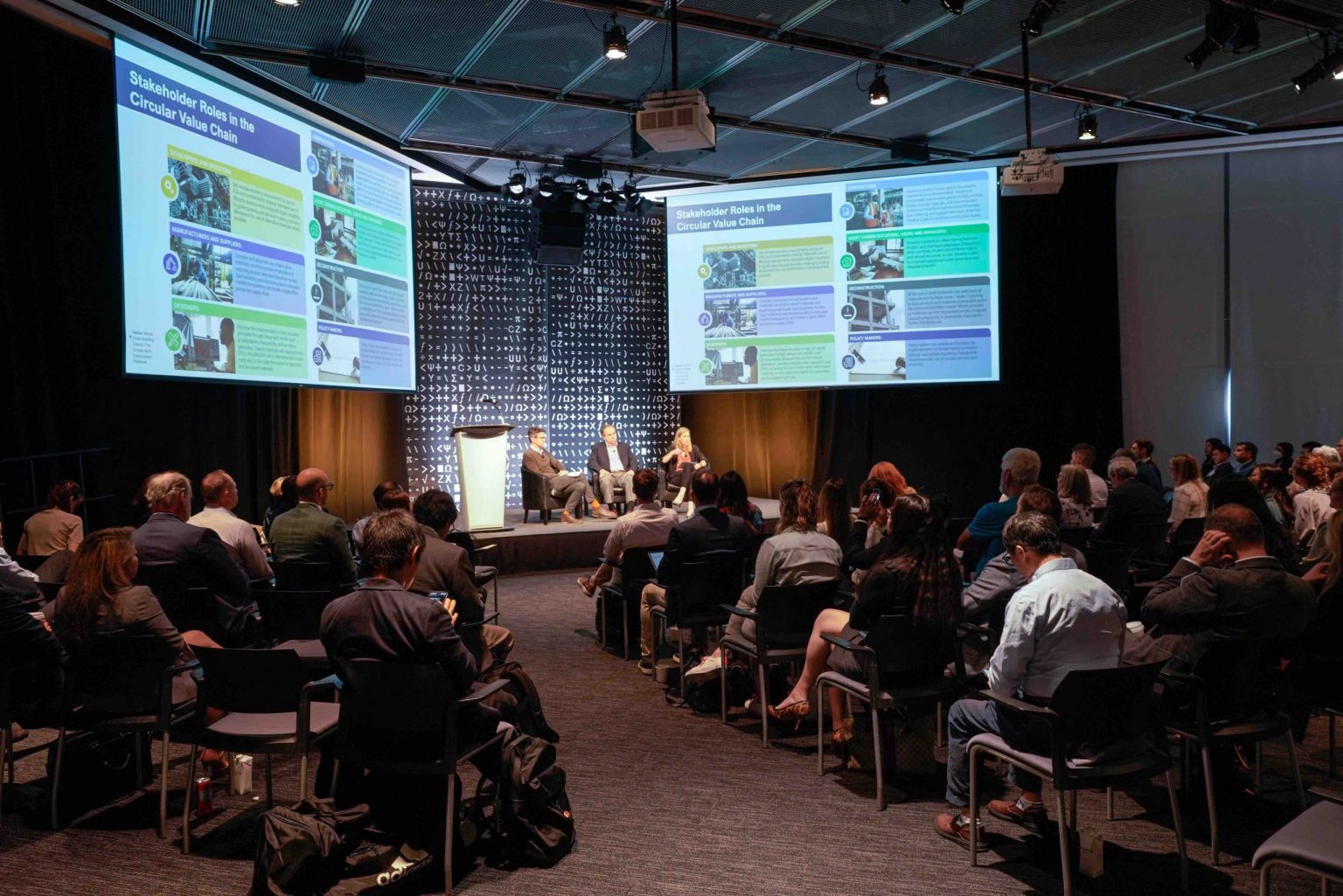 3 panelists sit on a dramatically lit stage with audience members surrounding, two screens project slides on either side of the stage depicting items and materials that can be reused in the building process